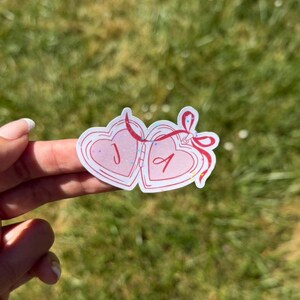 May include: A die-cut sticker featuring two connected heart-shaped lockets outlined in red. The left heart contains the letter J and the right heart contains the letter A in red script. A red ribbon bow connects the two hearts with holographic glitter.