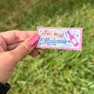 May include: A rectangular sticker with a scalloped edge featuring a holographic finish. The design includes illustrations of a pizza slice, a burger, and a wine bottle, with the text Malone's written in a blue script font surrounded by small stars.