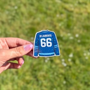 Pu&ograve; includere: Un adesivo blu a forma di maglia da hockey con il nome "Di Laurentis" e il numero "66". La maglia ha strisce bianche e azzurre sulle maniche e sul fondo. Lo sfondo &egrave; erba verde sfocata.
