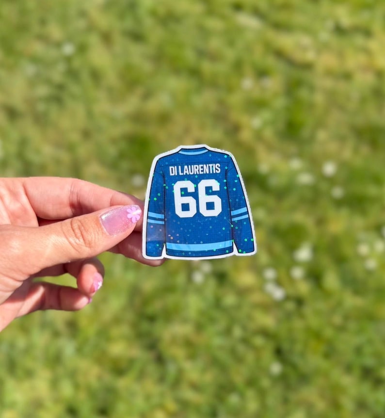 Pu&ograve; includere: Un adesivo blu a forma di maglia da hockey con il nome "Di Laurentis" e il numero "66". La maglia ha strisce bianche e azzurre sulle maniche e sul fondo. Lo sfondo &egrave; erba verde sfocata.