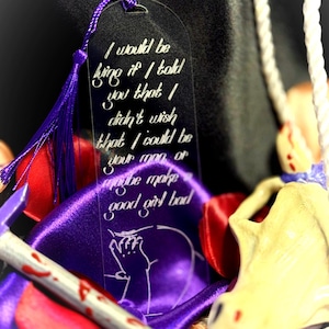 May include: A clear acrylic bookmark featuring a purple tassel and white engraved text that reads: I would be lying if I told you that I didn't wish that I could be your man or maybe make a good girl bad. Below the text is a line art illustration.