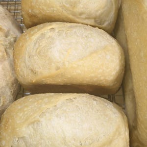 May include: Close-up of several loaves of freshly baked bread. The bread has a light golden-brown crust and a soft, airy interior. The loaves are rectangular and sit on a metal cooling rack. The image highlights the texture and color of the bread.