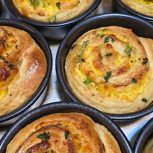 May include: Several golden-brown savory rolls baked in individual round black metal tins. Each spiral-shaped roll features a swirled dough texture with visible melted yellow cheese and flecks of green herbs throughout the baked surface.