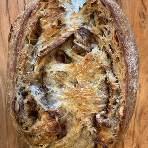 May include: Overhead shot of a rustic loaf of bread with a golden-brown crust and a textured, open crumb. The bread is oval-shaped and sits on a wooden surface, showcasing its artisanal quality. The crust has a slightly charred appearance.