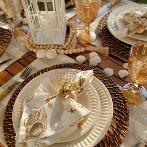 May include: A table setting with a white lantern, wooden beads, and a white napkin with a gold antler napkin ring. The table is set with a brown wicker charger, a white plate, and a gold-rimmed glass.
