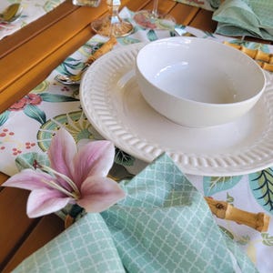May include: A table setting with a white plate and bowl, a green patterned napkin with a pink flower, and a wooden placemat.