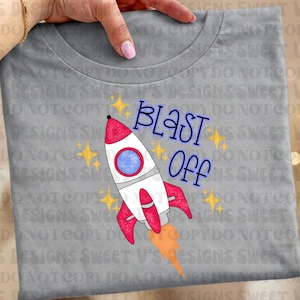 Puede incluir: Camiseta gris con un diseño de cohete colorido y las palabras "BLAST OFF" en azul. El cohete es blanco con detalles rojos y un ojo de buey azul, rodeado de estrellas amarillas.