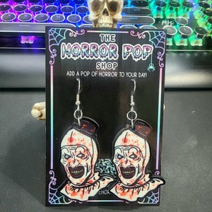 Könnte beinhalten: Ein Paar Hängeohrringe mit einem weißen und roten Clown-Gesicht mit schwarzem Umriss und roten Spritzern. Die Ohrringe befinden sich auf einem schwarzen Hintergrund mit einem weißen Spinnennetz-Design und dem Text "THE HORROR POP SHOP ADD A POP OF HORROR TO YOUR DAY!"