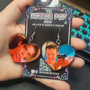 May include: A pair of heart-shaped earrings featuring images of characters from the film "American Psycho". The earrings are on a black card with the text "The Horror Pop Shop" and "Add a pop of horror to your day!" The card also includes a social media handle: @thehorrorpopshop.
