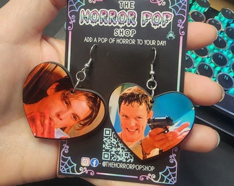 Könnte beinhalten: Ein Paar herzförmige Ohrringe mit Bildern von Charakteren aus dem Film "American Psycho". Die Ohrringe befinden sich auf einer schwarzen Karte mit dem Text "The Horror Pop Shop" und "Add a pop of horror to your day!" Die Karte enthält auch einen Social-Media-Handle: @thehorrorpopshop.
