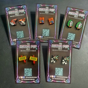 Peut inclure: Cinq cartes avec des bordures noires et du texte blanc qui dit "The Horror Pop Shop" et "Add a pop of horror to your day". Chaque carte présente une paire d'épingles en émail sur le thème des films d'horreur. Les épingles comprennent Beetlejuice,  Beetlejuice,  les sœurs Sanderson,  la  créature du lagon noir et  le  panneau Betelgeuse.