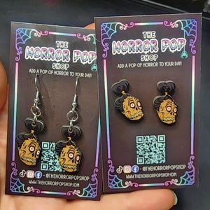 Könnte beinhalten: Zwei Paar Ohrringe mit einem Cartoon-Charakter mit einem Schädelgesicht und einem Dutt. Die Ohrringe befinden sich auf einer schwarz-weißen Karte mit dem Text "The Horror Pop Shop" und einem Spinnennetz-Design. Der Text "Add a pop of horror to your day!" ist ebenfalls auf der Karte.