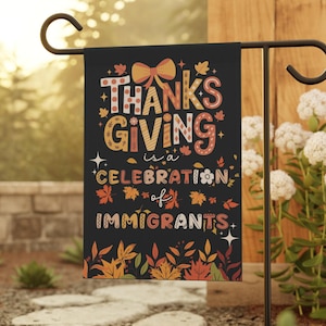 May include: A black garden flag with the text "Thanksgiving is a Celebration of Immigrants" in colorful lettering and fall-themed illustrations. The flag is displayed outdoors on a black metal stand, surrounded by greenery.