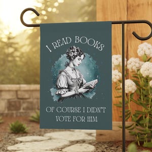 May include: A teal blue garden flag with a woman reading a book. The text on the flag reads "I read books of course I didn't vote for him."
