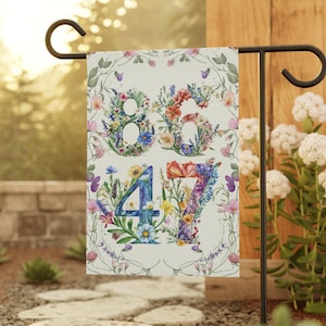 May include: A garden flag with the numbers 86 and 47 formed from colorful flowers and greenery on a white background.