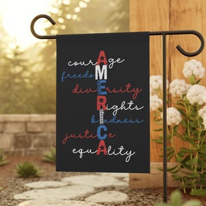 May include: A black garden flag with the word "AMERICA" in red, white, and blue, with words like "courage," "freedom," "diversity," "rights," "kindness," "justice," and "equality" written on it. The flag is displayed outdoors on a black metal stand.