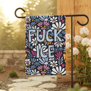 May include: A garden flag with the words "FUCK ICE" in blue and orange, surrounded by a floral pattern of white, pink, and blue flowers on a black background. The flag is displayed on a black metal stand.