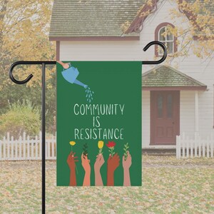 May include: A green garden flag with the text "Community is Resistance" in white. The flag features a blue watering can pouring water onto a row of hands holding flowers. The flowers are yellow, red, and orange.