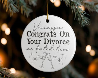 Puede incluir: Adorno navideño redondo de cerámica blanca con el texto "Vanessa, Congrats On Your Divorce we hated him." Tiene una cuerda dorada para colgar e ilustraciones de copas de champán y estrellas.