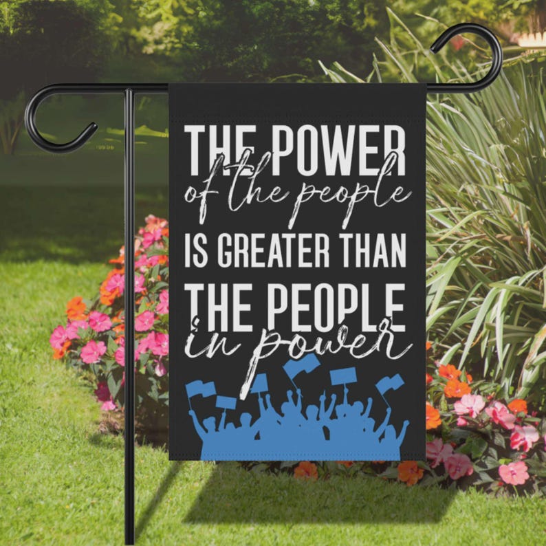 A black garden flag featuring the Cory Booker quote THE POWER OF THE PEOPLE IS GREATER THAN THE PEOPLE IN POWER in bold white lettering above a blue silhouette of a crowd of protesting citizens with signs and raised fists