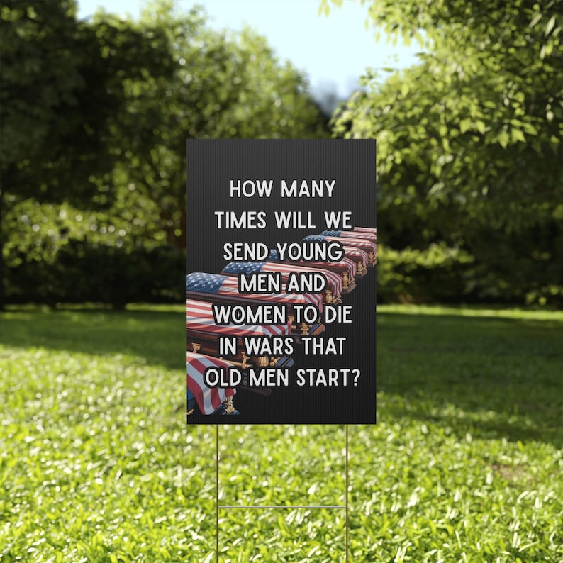 A black yard sign featuring a row of flag draped caskets behind the words HOW MANY TIMES WILL WE SEND YOUNG MEN AND WOMEN TO DIE IN WARS THAT OLD MEN START in bold white lettering
