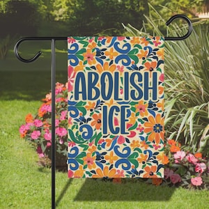 May include: A garden flag with the words "ABOLISH ICE" in blue, set against a colorful floral pattern of orange, yellow, and pink flowers on a cream background. The flag is displayed on a black metal stand.