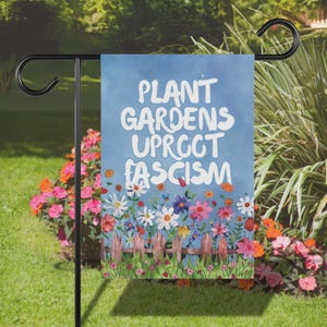 May include: A blue garden flag with white text that reads "Plant Gardens Uproot Fascism". The flag is decorated with colorful flowers and a picket fence.