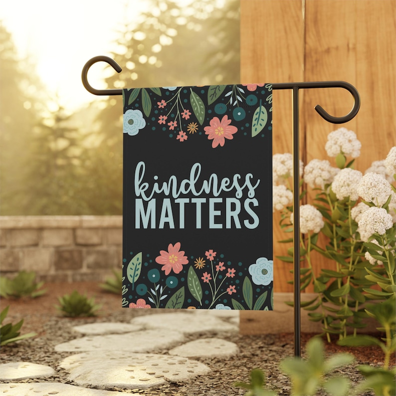 A black garden flag featuring a row of pastel flowers and greenery across the top and bottom framing the words KINDNESS MATTERS in light blue lettering in the center