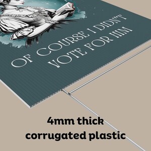 May include: A teal blue corrugated plastic yard sign with a black and white illustration of a woman reading a book. The text on the sign reads "Of course I didn't vote for him".