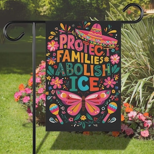 May include: Black garden flag with colorful text and illustrations. The flag reads "PROTECT FAMILIES ABOLISH ICE" with a sombrero, butterfly, flowers, and maracas. The flag is hanging on a black metal stand in a garden.