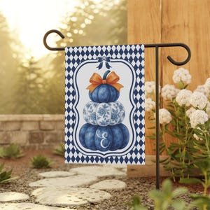 May include: A garden flag featuring a stack of three blue pumpkins with an orange bow on top. The middle pumpkin has a floral design. The flag has a white border with a blue diamond pattern and the letter "E".