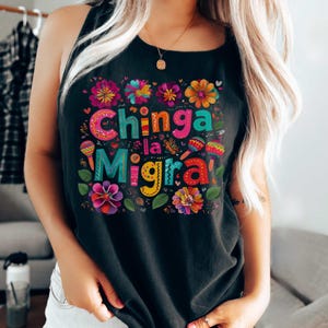 A woman wears a black tank top featuring a festive Mexican design with flowers and maracas surrounding the words CHINGA LA MIGRA
