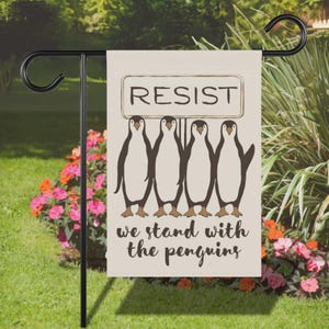 May include: A beige garden flag with a brown penguin illustration. The penguins are holding a sign that says "RESIST". The text below the penguins says "we stand with the penguins".