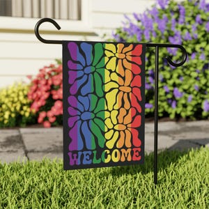May include: A black garden flag with a rainbow floral design and the word "WELCOME" in colorful letters. The flag is displayed on a black metal stand in a grassy yard, with colorful flowers in the background.