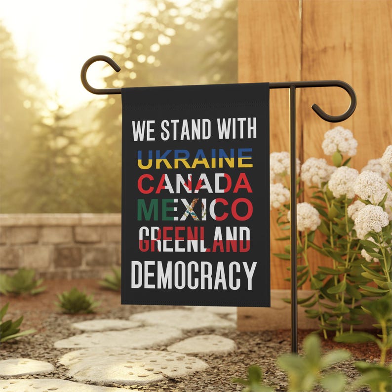 A black garden flag with the words We Stand With at the top in bold white lettering followed by Ukraine Canada Mexico Greenland in which every country name is shown in the color and pattern of their flag and the word Democracy at the bottom in bold white lettering