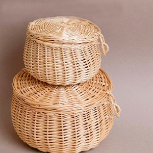 May include: Two woven wicker baskets, one stacked on top of the other. Each basket has a round shape with a matching lid. The baskets are a light tan colour and appear to be handmade.