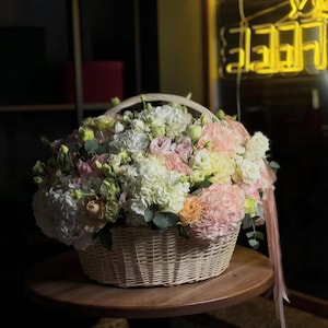 May include: A wicker basket overflowing with a variety of fresh flowers in shades of white, pink, peach, and yellow. The basket sits on a round wooden table, with a ribbon hanging from the side. The arrangement includes hydrangeas and roses.
