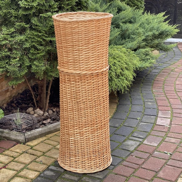 Könnte beinhalten: Ein hoher, zylindrischer Weidenkorb mit ausgestelltem Oberteil und gewebtem Design. Der Korb ist aus hellbraunem Weidengeflecht gefertigt und für den Außenbereich geeignet. Der Korb ist etwa 100 cm hoch.