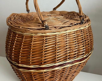 May include: A woven wicker basket with a rounded shape and a hinged lid. The basket features a sturdy handle for carrying. The basket is made of light brown wicker with a darker brown trim and accents.