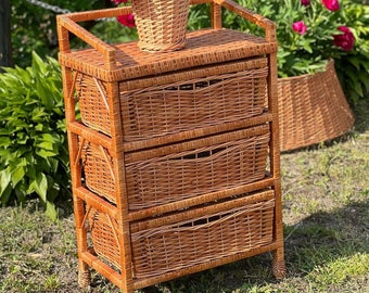May include: A three-drawer wicker storage unit with a natural brown finish. The unit is sitting on a green grassy surface with a wicker basket filled with pink flowers on top.