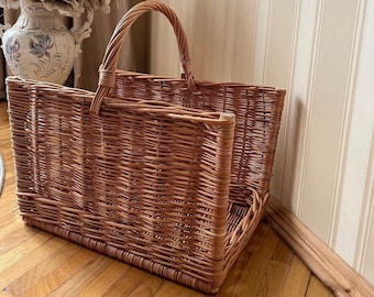May include: A woven wicker basket with a handle, perfect for storing firewood or other items. The basket is light brown and has a natural, rustic look.