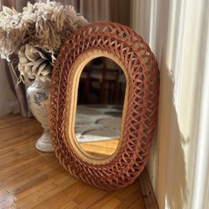 May include: Oval-shaped wicker mirror with a woven frame. The mirror has a natural brown colour and a light brown inner frame.