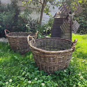 May include: Two large, round, woven wicker baskets with rope handles sit on a bed of green grass. A small wooden windmill and a tree are in the background, creating a rustic, outdoor scene.