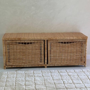Peut inclure: Un banc de rangement en osier de couleur naturelle avec deux tiroirs coulissants. Le banc est rectangulaire avec un motif tissé et un dessus plat. Les tiroirs ont une forme rectangulaire et une découpe de poignée. Le banc est posé sur un sol carrelé blanc.