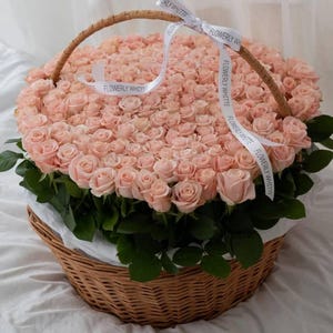 Puede incluir: Una cesta de mimbre rebosante de rosas de color rosa claro, acentuada con follaje verde y una cinta blanca. La cinta lleva la inscripción "FLOWERY WHITT". La cesta tiene un asa tejida.