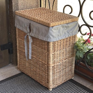 Puede incluir: Una cesta de lavandería rectangular de mimbre con un forro y tapa de tela gris claro. La cesta está hecha de mimbre tejido de color marrón claro y tiene una tapa a juego. El forro de tela es visible alrededor del borde superior, asegurado con una corbata gris claro.