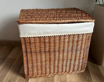 May include: A woven wicker laundry basket with a white fabric lid and a white lace trim. The basket is sitting on a wooden floor.