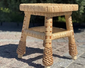 Pode incluir: Um pequeno banquinho de vime tecido natural com tampo quadrado e quatro pernas. As pernas são envoltas em um padrão tecido e têm um acabamento em madeira natural.