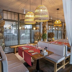 K&ouml;nnte beinhalten: Ein Restaurant-Essbereich mit wei&szlig;en, gepolsterten B&auml;nken und Holztischen. Die Tische sind mit rot-wei&szlig; karierten Tischdecken gedeckt und haben geflochtene Pendelleuchten dar&uuml;ber h&auml;ngen.