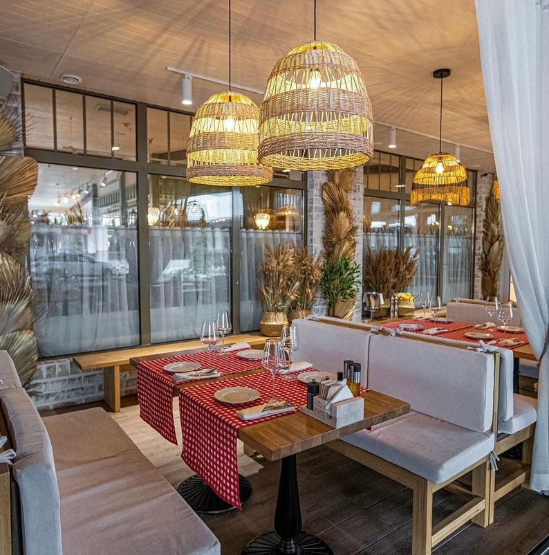 K&ouml;nnte beinhalten: Ein Restaurant-Essbereich mit wei&szlig;en, gepolsterten B&auml;nken und Holztischen. Die Tische sind mit rot-wei&szlig; karierten Tischdecken gedeckt und haben geflochtene Pendelleuchten dar&uuml;ber h&auml;ngen.